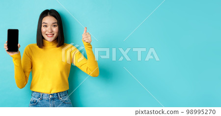 Technology and people concept. Cheerful asian girl in yellow sweater showing blank smartphone screen and thumbs up, demonstrate online offer, standing over blue background 98995270