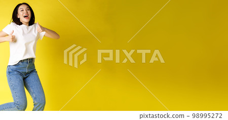Full size of happy young asian woman jumping from joy, showing thumbs up in approval, posing over yellow background in jeans and casual white t-shirt Full size of happy young asian woman jumping from joy, showing thumbs up in approval, posing over yellow background in jeans and casual white t-shirt 98995272