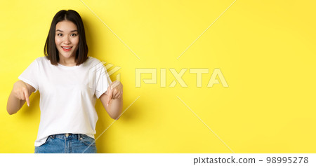 Beauty and fashion concept. Beautiful asian woman in white t-shirt pointing fingers down, demonstrate logo standing over yellow background 98995278