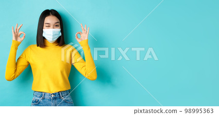 Covid-19, social distancing and pandemic concept. Cheerful asian girl in medical mask showing okay gestures, winking at camera, showing approval, standing over blue background Covid-19, social distancing and pandemic concept. Cheerful asian girl in medical mask showing okay gestures, winking at camera, showing approval, standing over blue background 98995363