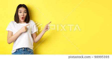 Beauty and fashion concept. Beautiful asian woman in white t-shirt pointing fingers right, demonstrate logo standing over yellow background 98995393
