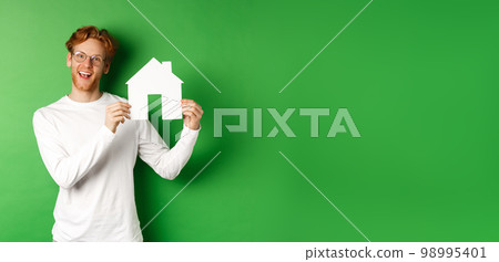 Real estate and buying property concept. Handsome young man with red hair showing house cutout, searching for new home, standing over green background 98995401