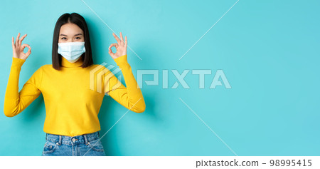 Covid-19, social distancing and pandemic concept. Smiling confident asian woman in medical mask showing OK signs, approve or praise good deal, standing over blue background Covid-19, social distancing and pandemic concept. Smiling confident asian woman in medical mask showing OK signs, approve or praise good deal, standing over blue background 98995415