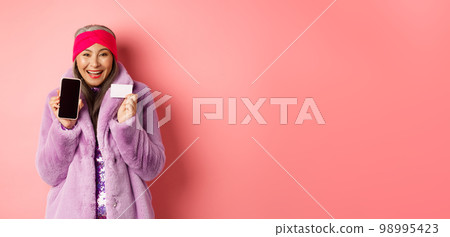 Online shopping and fashion concept. Beautiful asian mature woman showing plastic credit card and blank phone screen, smiling happy, pink background 98995423