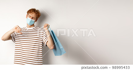 Concept of social distancing and shopping. Disappointed young man with red hair, wearing face mask, holding shopping bag and showing thumb-down from dislike, disapprove store 98995433