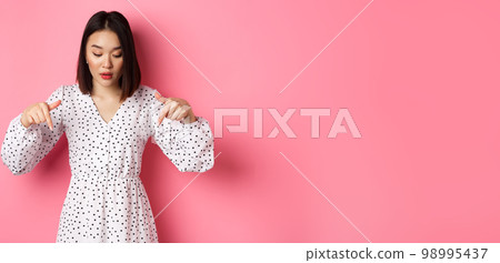 Romantic asian woman peeking down, pointing fingers at bottom, looking curious at product discount, standing over pink background 98995437