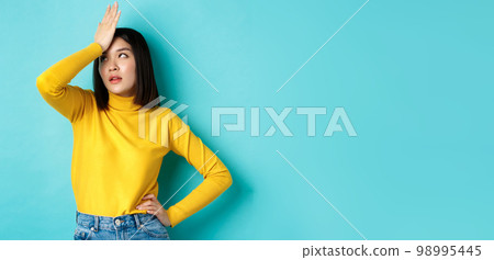 Emotions and lifestyle concept. Annoyed asian girl roll eyes and face palm, standing bothered in yellow pullover against blue background 98995445