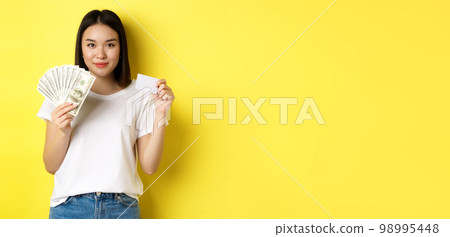 Beautiful asian woman with short dark hair, wearing white t-shirt, showing money in dollars and plastic credit card, standing over yellow background Beautiful asian woman with short dark hair, wearing white t-shirt, showing money in dollars and plastic credit card, standing over yellow background 98995448