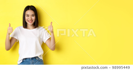 Pretty young asian woman in white t-shirt, showing thumbs up and smiling, praise good offer, recommend product, standing satisfied over yellow background Pretty young asian woman in white t-shirt, showing thumbs up and smiling, praise good offer, recommend product, standing satisfied over yellow background 98995449