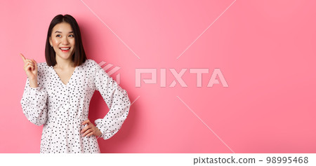 Happy asian girl in dress smiling satisfied, pointing finger at upper left corner, nod in approval, standing in dress against pink background 98995468
