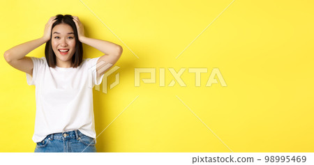 Amazed asian girl winning prize and looking with excitement and disbelief, holding hands on head and smiling happy, standing over yellow background 98995469