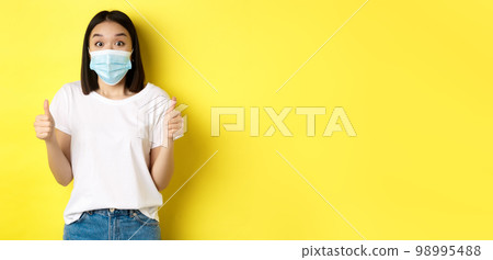 Covid-19, quarantine and social distancing concept. Cheerful asian woman in medical mask and white t-shirt showing thumbs up in approval, praise good deal, yellow background Covid-19, quarantine and social distancing concept. Cheerful asian woman in medical mask and white t-shirt showing thumbs up in approval, praise good deal, yellow background 98995488