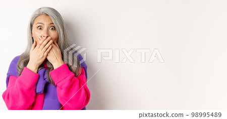 Close up of shocked senior lady gasping and covering mouth, staring at camera speechless, witness something, standing over white background Close up of shocked senior lady gasping and covering mouth, staring at camera speechless, witness something, standing over white background 98995489