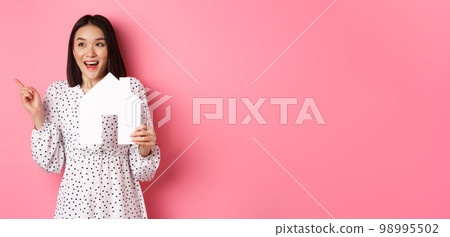 Real estate. Excited asian woman showing paper house model, pointing and looking left at copy space, standing over pink background 98995502