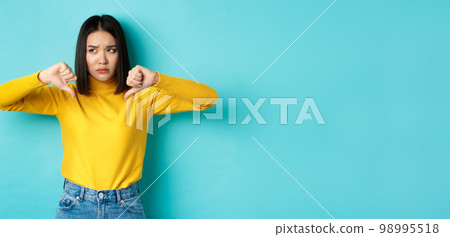 Disappointed asian woman frowning upset, showing thumbs down in dislike and disapproval, standing over blue background, looking left Disappointed asian woman frowning upset, showing thumbs down in dislike and disapproval, standing over blue background, looking left 98995518