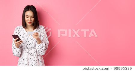 Beautiful asian woman enter credit card info in mobile phone app, shopping online, paying for order on smartphone, standing over pink background 98995559