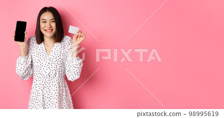 Joyful korean woman showing smartphone screen and credit card, paying for internet order, demonstrating online shopping app, standing over pink background Joyful korean woman showing smartphone screen and credit card, paying for internet order, demonstrating online shopping app, standing over pink background 98995619