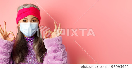 Covid-19, virus and fashion concept. Close-up of fashionable asian senior woman in face mask, showing OK signs and looking happy, pink background Covid-19, virus and fashion concept. Close-up of fashionable asian senior woman in face mask, showing OK signs and looking happy, pink background 98995621