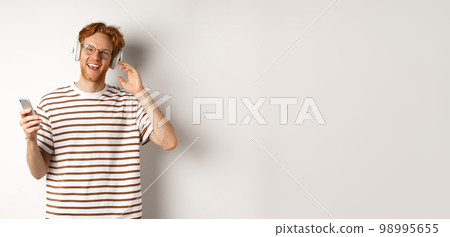 Technology concept. Young man with red hair and beard listening music in headphones and using smartphone, smiling at camera, white background Technology concept. Young man with red hair and beard listening music in headphones and using smartphone, smiling at camera, white background 98995655