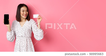 Happy young asian woman internet shopping, showing smartphone screen and looking satisfied at credit card, standing over pink background Happy young asian woman internet shopping, showing smartphone screen and looking satisfied at credit card, standing over pink background 98995663