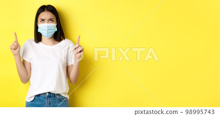 Covid-19, pandemic and social distancing concept. Disappointed asian girl in medical mask, frowning upset and pointing fingers up at logo, standing over yellow background Covid-19, pandemic and social distancing concept. Disappointed asian girl in medical mask, frowning upset and pointing fingers up at logo, standing over yellow background 98995743