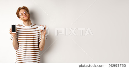 Shopping and finance concept. Pleased young man with red hair smiling from satisfaction, showing smartphone blank screen and credit card, white background 98995760