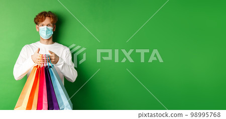Pandemic and lifestyle concept. Cheerful redhead man going shopping in store, wearing medical mask and holding bags, standing over green background Pandemic and lifestyle concept. Cheerful redhead man going shopping in store, wearing medical mask and holding bags, standing over green background 98995768