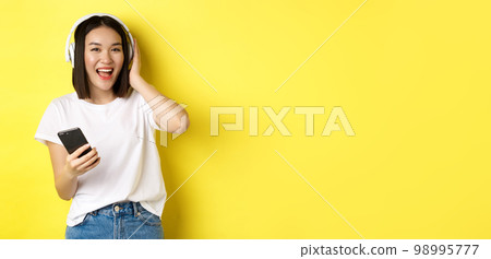 Cool asian girl dancing and listening music in wireless headphones, holding smartphone in hand, standing over yellow background 98995777
