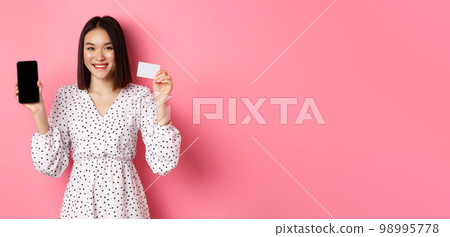 Cute asian woman shopping online, showing bank credit card and mobile screen, smiling and looking at camera, standing over pink background Cute asian woman shopping online, showing bank credit card and mobile screen, smiling and looking at camera, standing over pink background 98995778