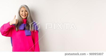 Portrait of happy and satisfied asian senior woman showing thumb-up, approval and like something, smiling and praising product, standing over white background Portrait of happy and satisfied asian senior woman showing thumb-up, approval and like something, smiling and praising product, standing over white background 98995807