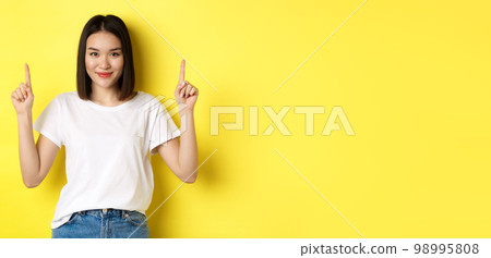 Beauty and fashion concept. Beautiful asian woman in white t-shirt pointing fingers up, standing over yellow background 98995808