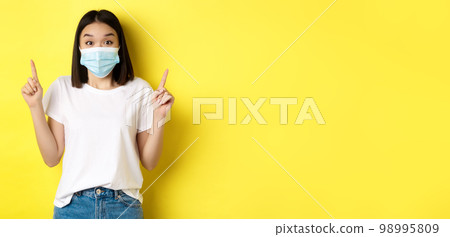 Covid-19, pandemic and social distancing concept. Young asian woman in white t-shirt and medical mask from coronavirus, pointing fingers up, showing special offer Covid-19, pandemic and social distancing concept. Young asian woman in white t-shirt and medical mask from coronavirus, pointing fingers up, showing special offer 98995809