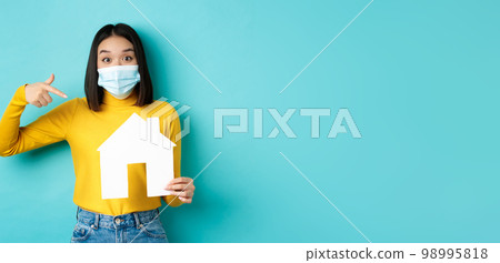 Covid-19, pandemic and real estate concept. Cheerful asian woman smiling in medical mask, showing paper house cutout, recommend agency for buying property, blue background Covid-19, pandemic and real estate concept. Cheerful asian woman smiling in medical mask, showing paper house cutout, recommend agency for buying property, blue background 98995818