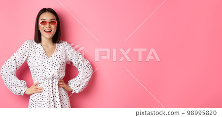 Tourism and lifestyle concept. Beautiful korean woman on vacation, wearing sunglasses and dress, looking left with happy smile, standing over pink background 98995820