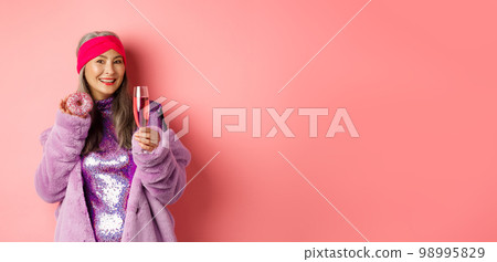 Elegant and stylish asian woman in purple faux fur coat eating donuts and drinking champagne, having fun at party, standing over pink background 98995829