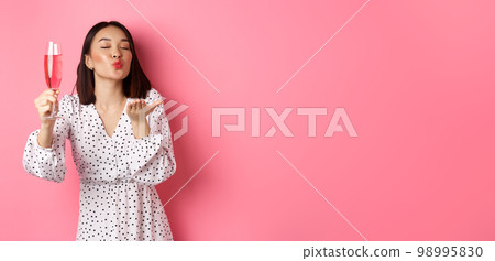 Romantic asian woman raising glass of champagne and sending air kiss at camera, celebrating and having fun, standing over pink background 98995830