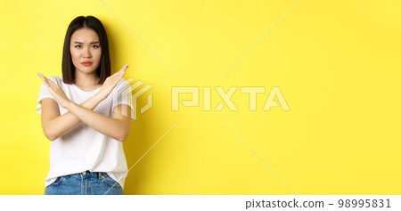 Serious and assertive korean woman showing cross stop gesture, frowning and telling no, prohibit action, disapprove something bad, standing over yellow background 98995831