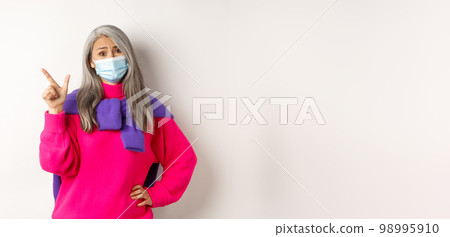 Covid-19, quarantine and lockdown concept. Skeptical asian elderly female in face mask looking disappointed, pointing at upper left corner, white background Covid-19, quarantine and lockdown concept. Skeptical asian elderly female in face mask looking disappointed, pointing at upper left corner, white background 98995910