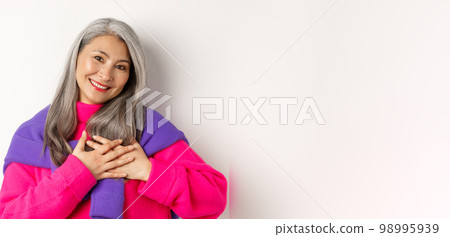 Close up of stylish asian mother holding hands on heart, looking touched and heartfelth, saying thank you, standing over white background 98995939