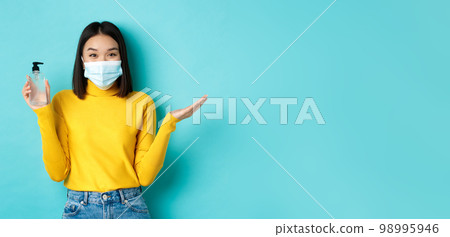 Covid-19, social distancing and pandemic concept. Cheerful asian woman showing hand sanitizer, recommend use antiseptic from coronavirus, wearing medical mask Covid-19, social distancing and pandemic concept. Cheerful asian woman showing hand sanitizer, recommend use antiseptic from coronavirus, wearing medical mask 98995946