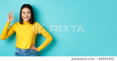 Happy beautiful asian woman smiling, showing okay gesture in approval, saying yes, standing pleased over blue background 98995960