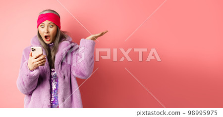 Online shopping and fashion concept. Amazed and surprised asian senior woman looking at smartphone screen, reading on mobile phone and gasping, pink background Online shopping and fashion concept. Amazed and surprised asian senior woman looking at smartphone screen, reading on mobile phone and gasping, pink background 98995975