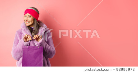 Shopping and fashion concept. Fashionable gradmother bought present and smiling, holding purple bag in hands, standing over pink background Shopping and fashion concept. Fashionable gradmother bought present and smiling, holding purple bag in hands, standing over pink background 98995978