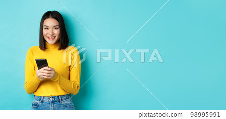 Beautiful and stylish asian woman shopping online on mobile phone, standing over blue background Beautiful and stylish asian woman shopping online on mobile phone, standing over blue background 98995991