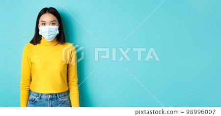 Covid-19, social distancing and pandemic concept. Teenage asian woman in medical mask, looking at camera and standing against blue background in yellow sweater 98996007