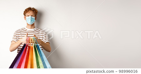 Pandemic and lifestyle concept. Cheerful redhead man going shopping in store, wearing medical mask and holding bags, standing over white background 98996016