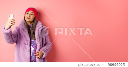 Fashion concept. Stylish asian senior female taking selfie on smartphone, posing in purple faux fur coat and party dress, standing over pink background Fashion concept. Stylish asian senior female taking selfie on smartphone, posing in purple faux fur coat and party dress, standing over pink background 98996025