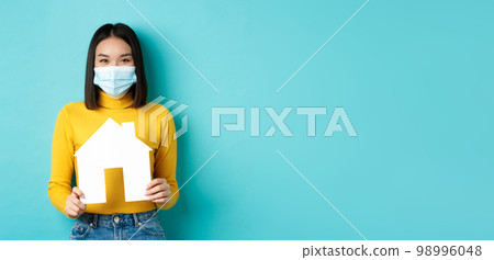 Covid-19, pandemic and real estate concept. Cheerful asian woman smiling in medical mask, showing paper house cutout, recommend agency for buying property, blue background Covid-19, pandemic and real estate concept. Cheerful asian woman smiling in medical mask, showing paper house cutout, recommend agency for buying property, blue background 98996048