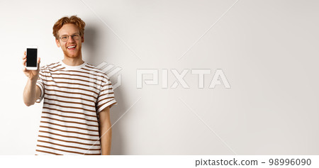 Technology and e-commerce concept. Happy young redhead man in glasses showing blank smartphone screen, smiling satisfied, standing over white background Technology and e-commerce concept. Happy young redhead man in glasses showing blank smartphone screen, smiling satisfied, standing over white background 98996090