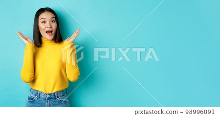 Beauty and fashion concept. Image of excited and surprised japanese girl saying wow with amazement, raising hands up near face, standing against blue background Beauty and fashion concept. Image of excited and surprised japanese girl saying wow with amazement, raising hands up near face, standing against blue background 98996091